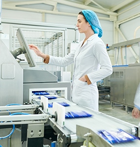 Busy Female Worker In Sterile Clothes Choosing Program On Touch Screen While Operating Manufacturing Machine Producing Packaged Food, Blurred Motion Of Technologists